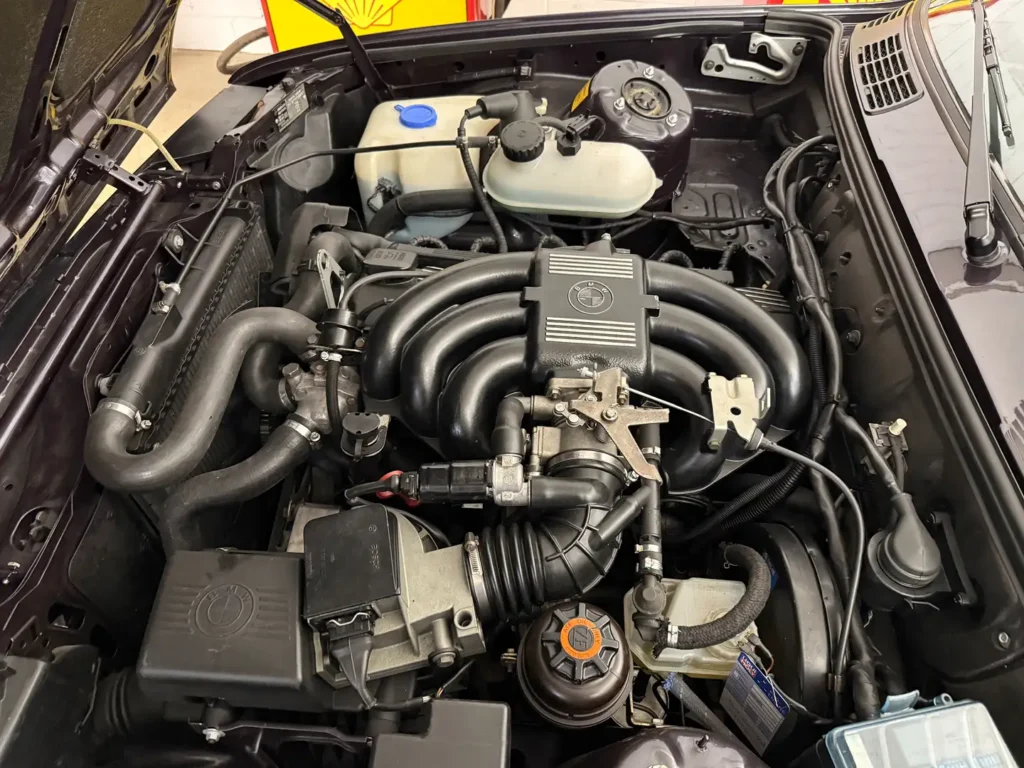 Offene Motorhaube eines BMW 325i E30 AC Schnitzer mit Blick auf den Motorraum, inklusive Motor und sämtlicher Anbauteile im Detail