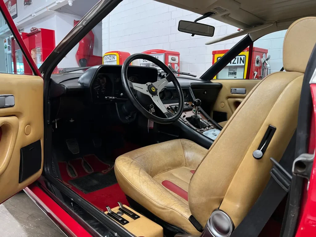 Innenraum eines Ferrari 400i mit beigen Ledersitzen, Lederlenkrad und Dachhimmel, dargestellt bei geöffneter Tür im klassischen Design
