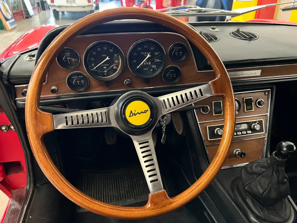 nnenraum eines Fiat Dino Spider mit Holzlenkrad, Schaltkonsole, Armaturenbrett, Radio und klassischen Bedienelementen im Fokus.